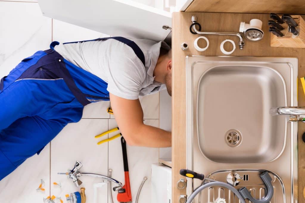 A plumber in blue workwear repairs a kitchen sink, with plumbing tools, pipes, fittings, and a faucet spread around the work area.