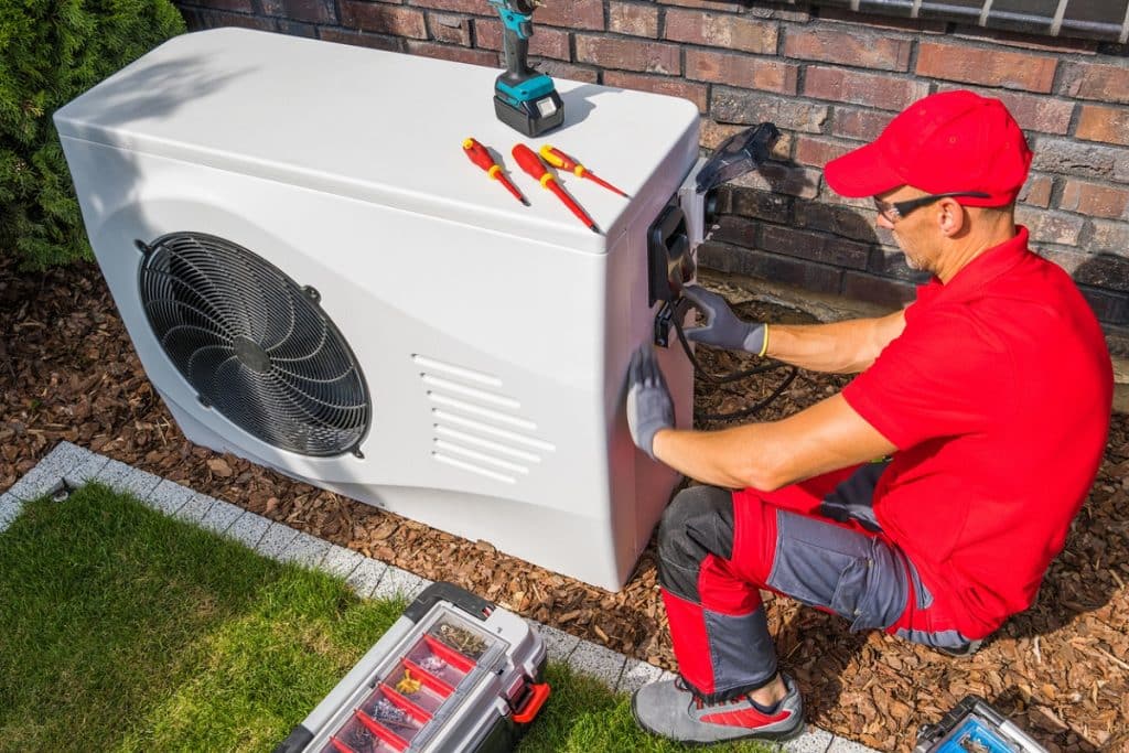 A technician in a red uniform repairs an outdoor heat pump unit using hand tools, with a toolbox and power drill nearby.
